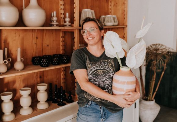 Eine lächelnde Frau steht mit einer Blumenvase im Arm vor einem Holzregal mit weiteren Vasen.